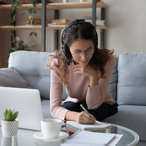 Junge Frau sitzt auf der Couch vor Laptop und schreibt etwas in einen Notizblock. Junge Frau sitzt auf der Couch vor Laptop und schreibt etwas in einen Notizblock.