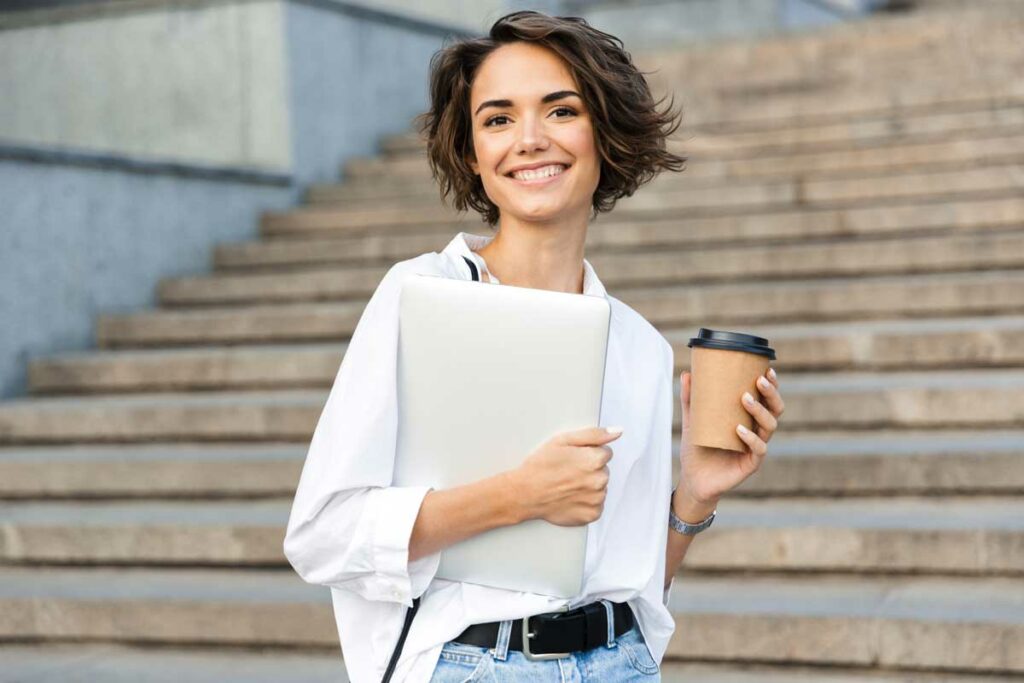 Junge Frau mit Laptop und Kaffee in der Hand steht auf einer Stiege Junge Frau mit Laptop und Kaffee in der Hand steht auf einer Stiege