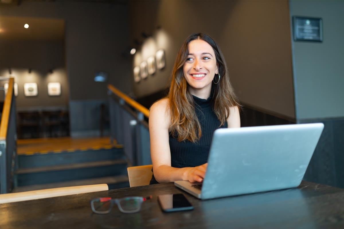 Frau sitzt mit Laptop im Restaurant und sieht lachend zur Seite.