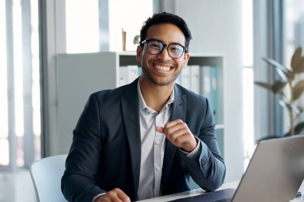 Lächelnder Mann im Business-Outfit sitzt an einem Schreibtisch mit Laptop in modernem Büro.