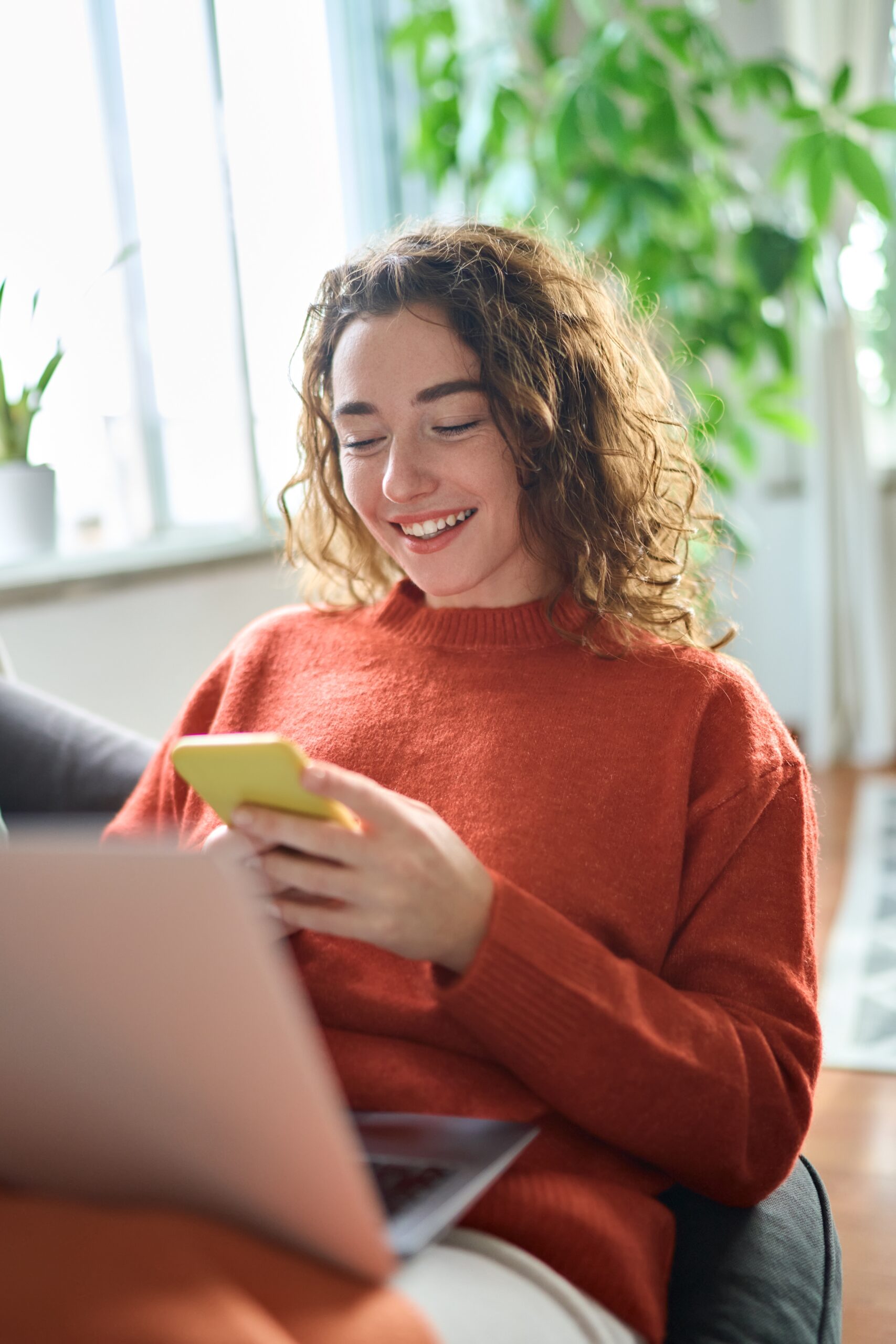 Junge Frau in rotem Pullover mit Locken, sitzt auf dem Sofa mit Laptop im Schoß und Handy in der Hand, lächelt
